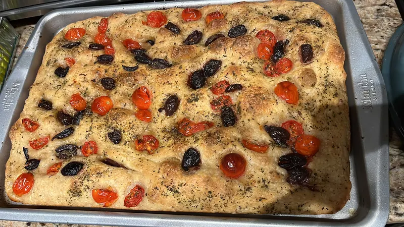 Sa focaccia dans la plaque, piquée de creux et garnie d’olives noires, de tomates cerises et d’herbes.